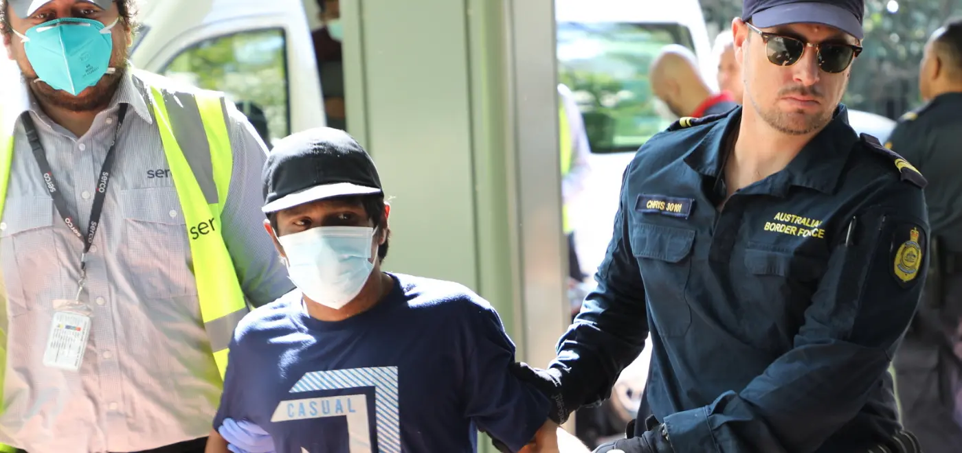 Petugas Australian Border Force mengawal seorang pria yang ditahan yang mengenakan masker dan pakaian kasual.